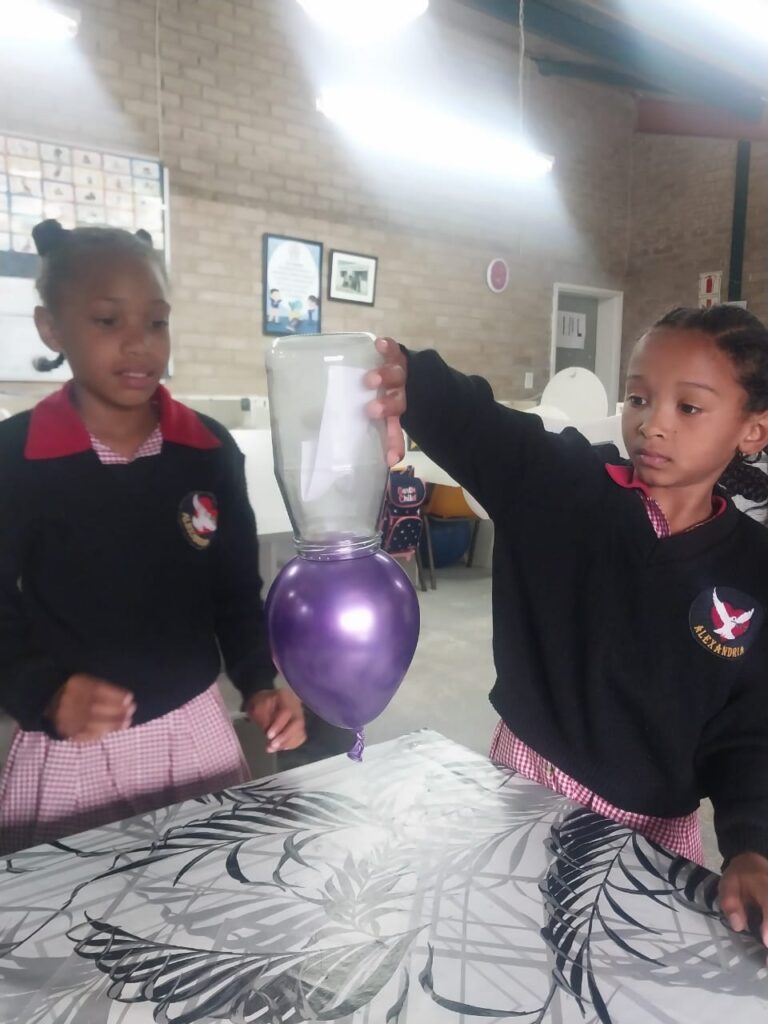 Miley Cannon demonstrates a scientific experiment to Leyana van Rooyen, encouraging curiosity and hands-on learning.